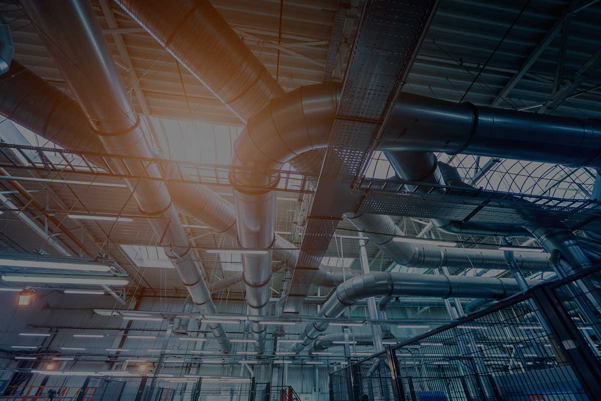Large metal ventilation ducts run across the ceiling of an industrial facility, illuminated by bright overhead lights, with steel beams and grates visible throughout the modern, spacious interior.