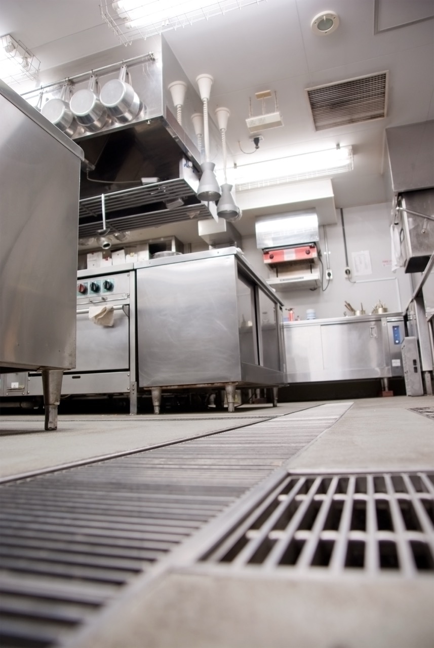 A commercial kitchen with stainless steel appliances and counters, various cooking equipment, pots hanging above, and ventilation grates on the floor—showcasing an ideal space maintained by professional restaurant cleaning services.