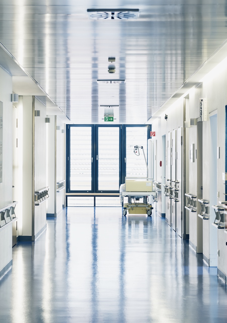 A clean, empty hospital hallway with shiny floors showcases the results of professional hospital cleaning services. White walls, closed doors on both sides, and a hospital bed near large windows are brightened by natural light filling the space.