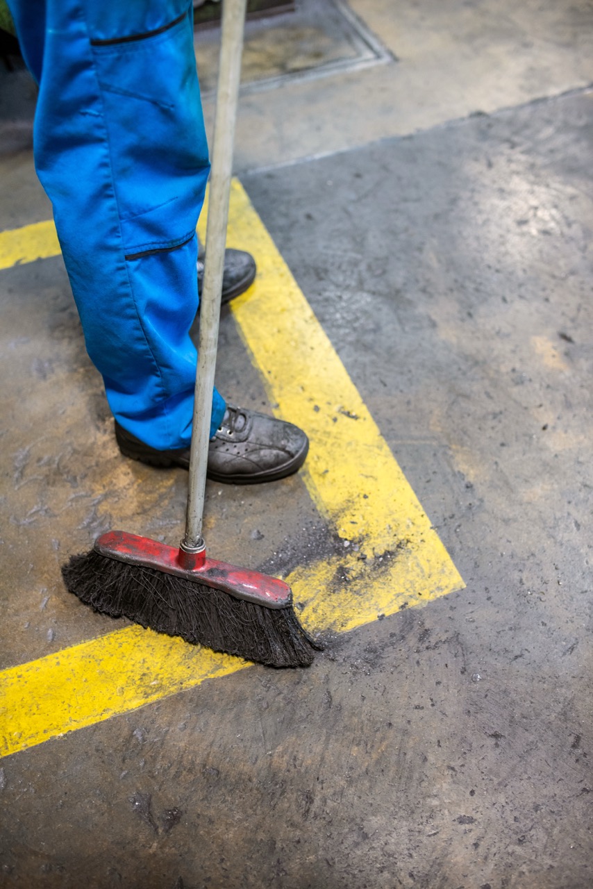 A person in blue pants and work shoes is sweeping a dirty industrial floor with a broom, cleaning near a yellow painted line.