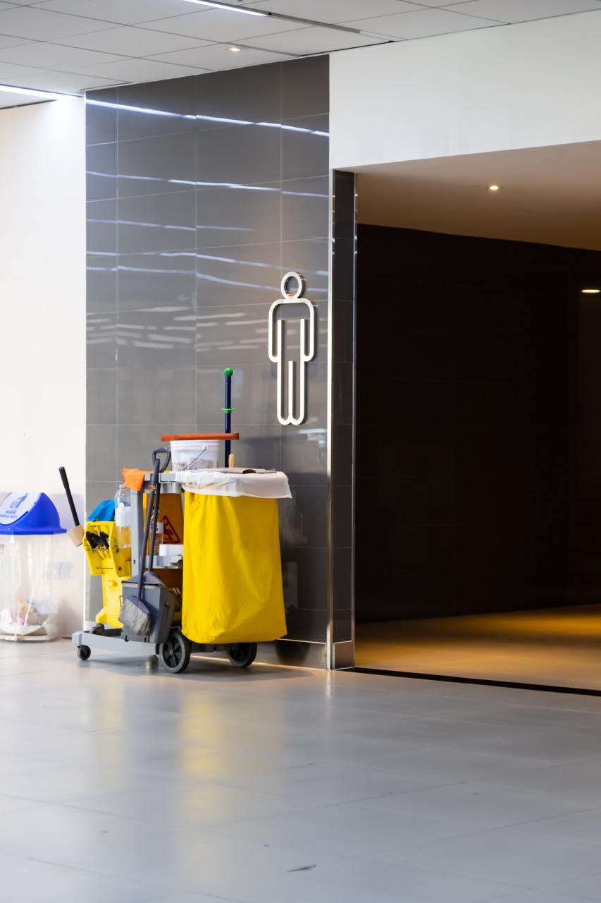 A janitor cart with cleaning supplies is parked next to a men's restroom entrance in an office building, identified by a white male figure sign on the tiled wall. The clean, well-lit surroundings reflect professional office building cleaning services.