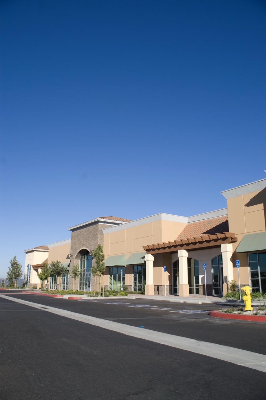 A row of empty storefronts in a modern shopping center with large windows and awnings, clear blue sky above, and an empty parking lot—ideal for shopping center cleaning services to keep spaces pristine.