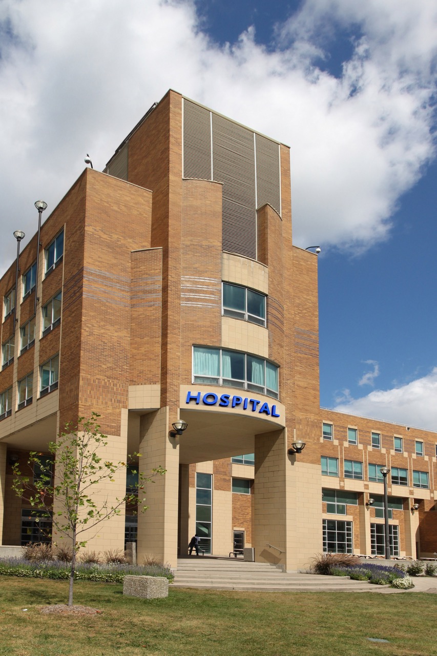 A modern, multi-story brick building with large windows and a blue "HOSPITAL" sign above the entrance, set against a partly cloudy sky. The hospital’s pristine appearance highlights quality hospital cleaning services, with greenery and a small tree in front.
