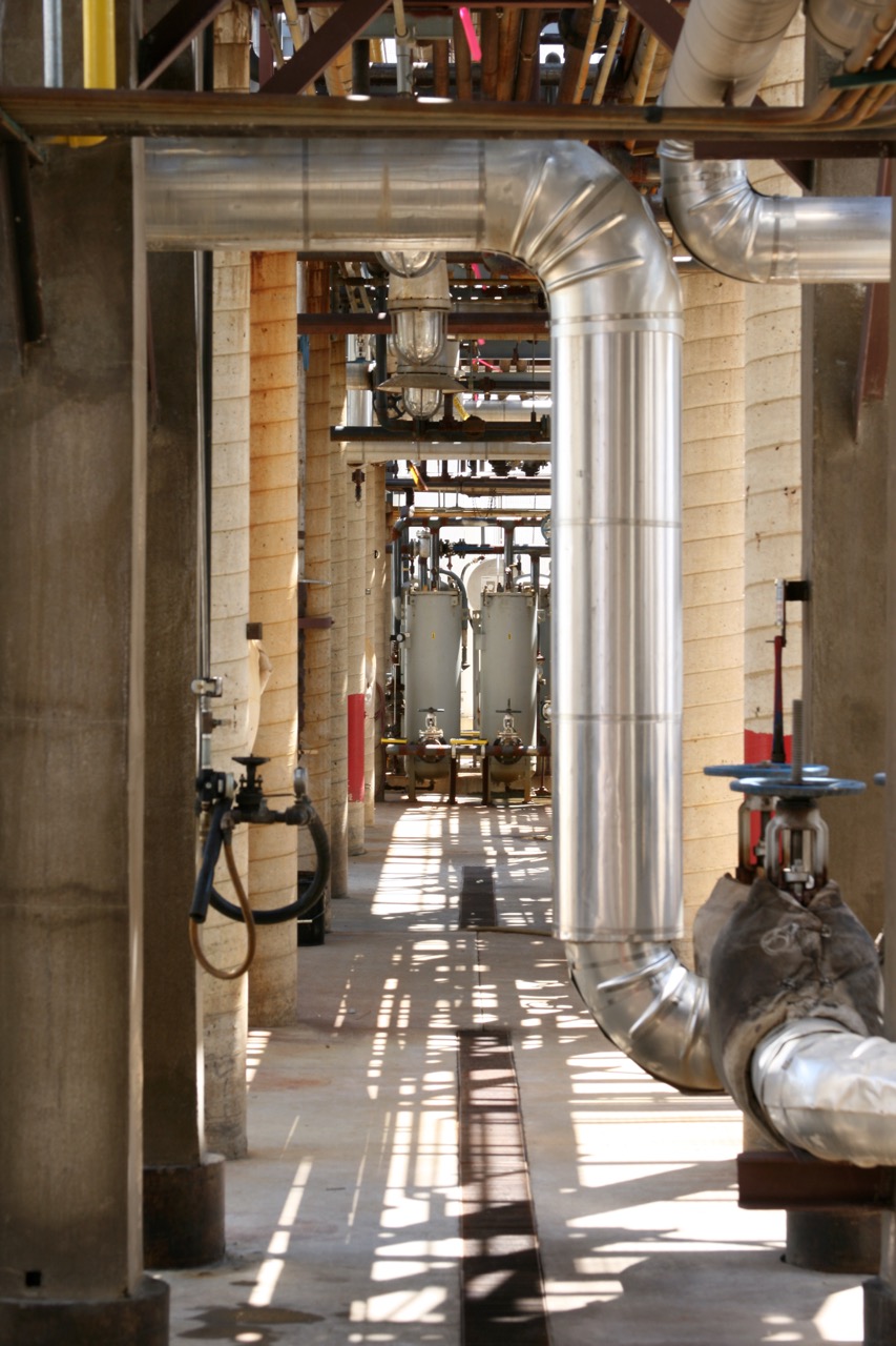 Industrial facility interior with large metal pipes, tanks, and concrete pillars. Sunlight casts shadows on the floor, highlighting the structure’s linear perspective and mechanical components—ideal for specialized oil and gas cleaning services.