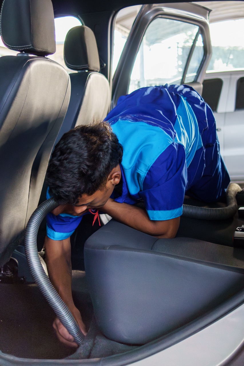 A person in a blue uniform is vacuuming the interior of a car, reaching under the front seat with a vacuum hose as part of professional fleet cleaning services.