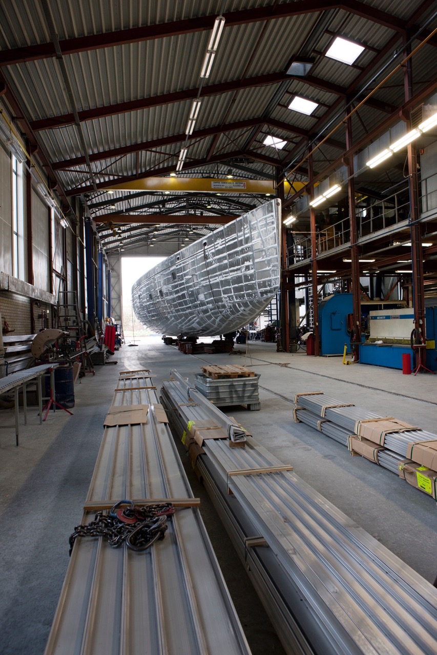 A large metal ship hull under construction sits in the middle of a spacious industrial workshop, surrounded by long metal beams, tools, and equipment. The brightly lit space with high ceilings is maintained to military cleaning services standards.