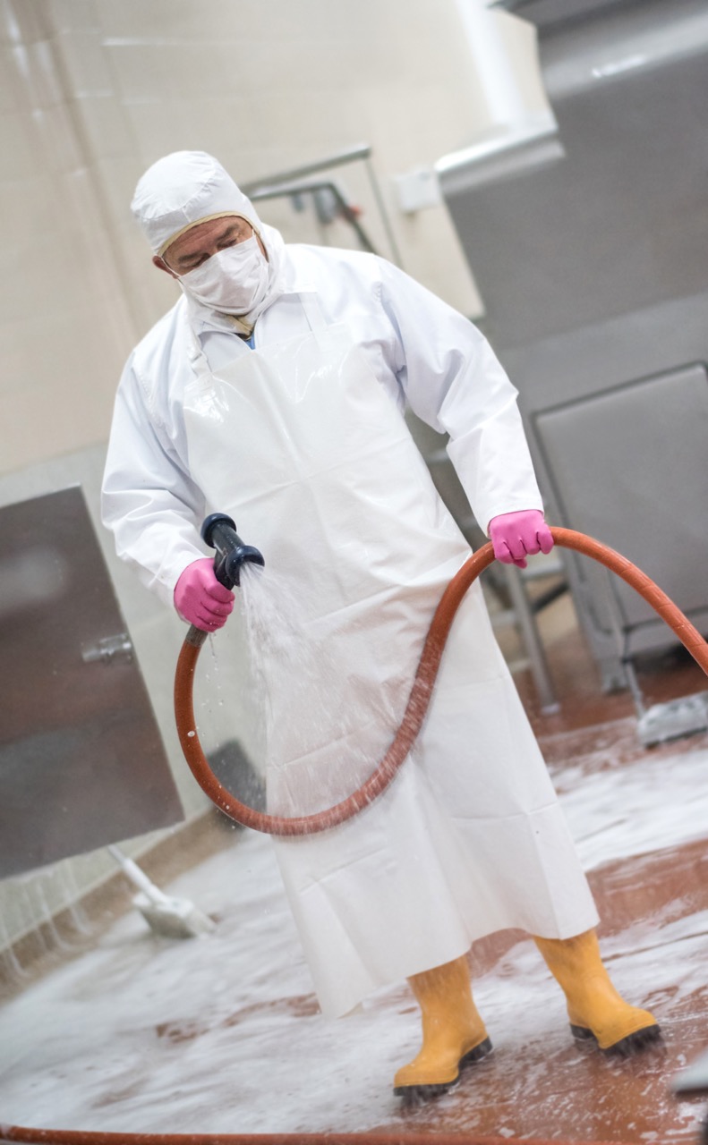 A worker in protective clothing, including a white gown, mask, gloves, and yellow boots, uses a hose to spray and clean a factory floor covered in foam—similar to restaurant cleaning services for maintaining hygiene.