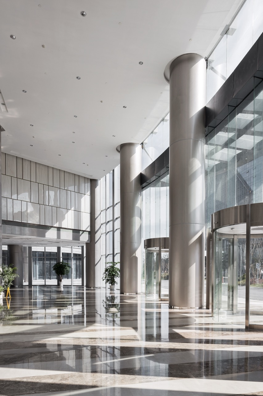 Spacious, modern lobby with tall glass windows, large cylindrical columns, glossy marble floors maintained by office building cleaning services, and some potted plants. Sunlight streams in, creating bright reflections and shadows across the space.