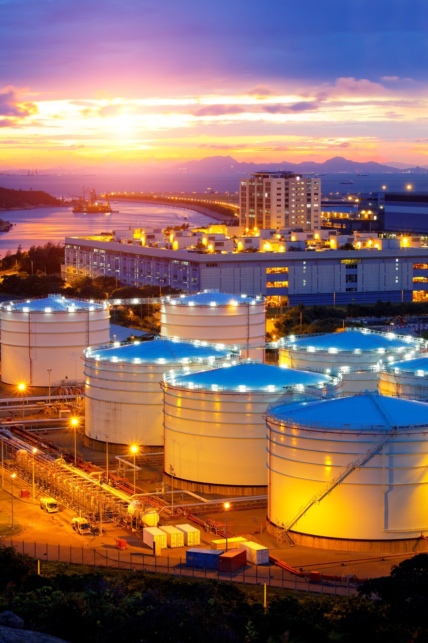 Large cylindrical storage tanks at an industrial facility, maintained with oil and gas cleaning services, are illuminated at sunset, with buildings, water, and mountains visible in the background under a colorful sky.