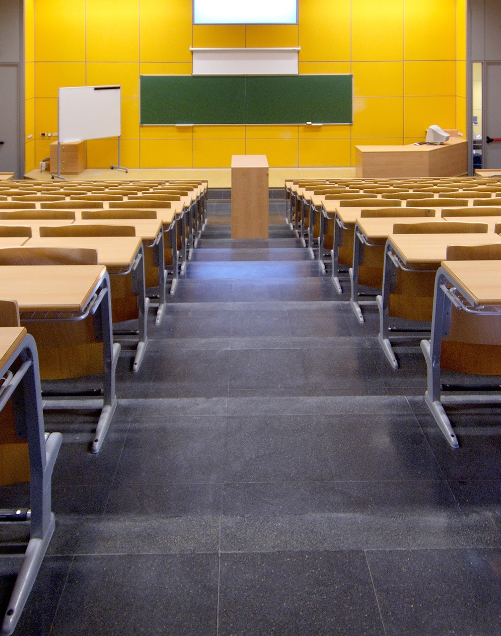 An empty lecture hall with rows of wooden seats, a central podium, green chalkboard, whiteboard, and projector screen on a yellow-paneled wall—spotless thanks to expert university cleaning services. The room is well-lit and inviting.