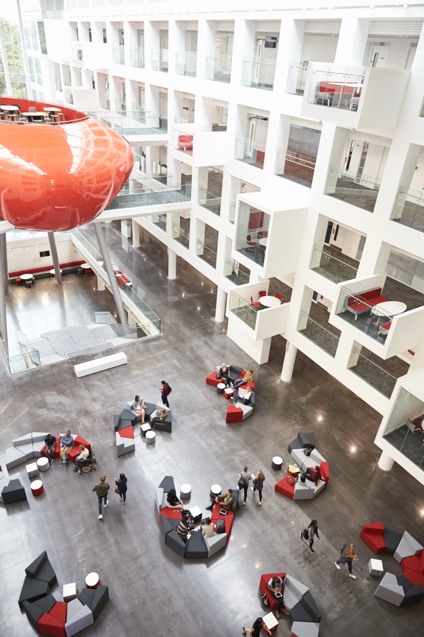 A modern, open atrium with geometric seating areas, groups of people talking, and an eye-catching red pod structure suspended above. White walls with cubed balconies line the upper levels, kept pristine by efficient university cleaning services.