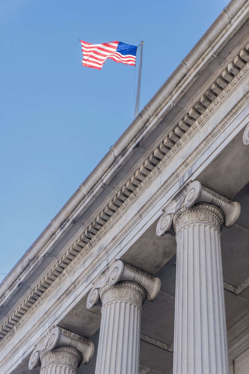 A U.S. flag waves atop a neoclassical building with tall, fluted columns and ornate capitals, maintained with expert government cleaning services, set against a clear blue sky.