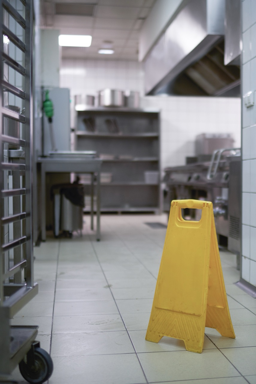 A yellow caution wet floor sign stands on the tiled floor of a commercial kitchen, where stainless steel shelves and equipment shine—highlighting the importance of restaurant cleaning services in maintaining safety and hygiene.