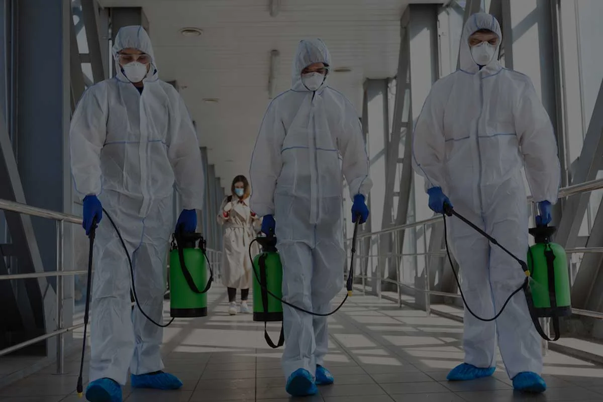 Three people in full-body protective suits, masks, and gloves hold green disinfectant sprayers while walking in a corridor. A fourth person in a white coat and mask stands in the background.