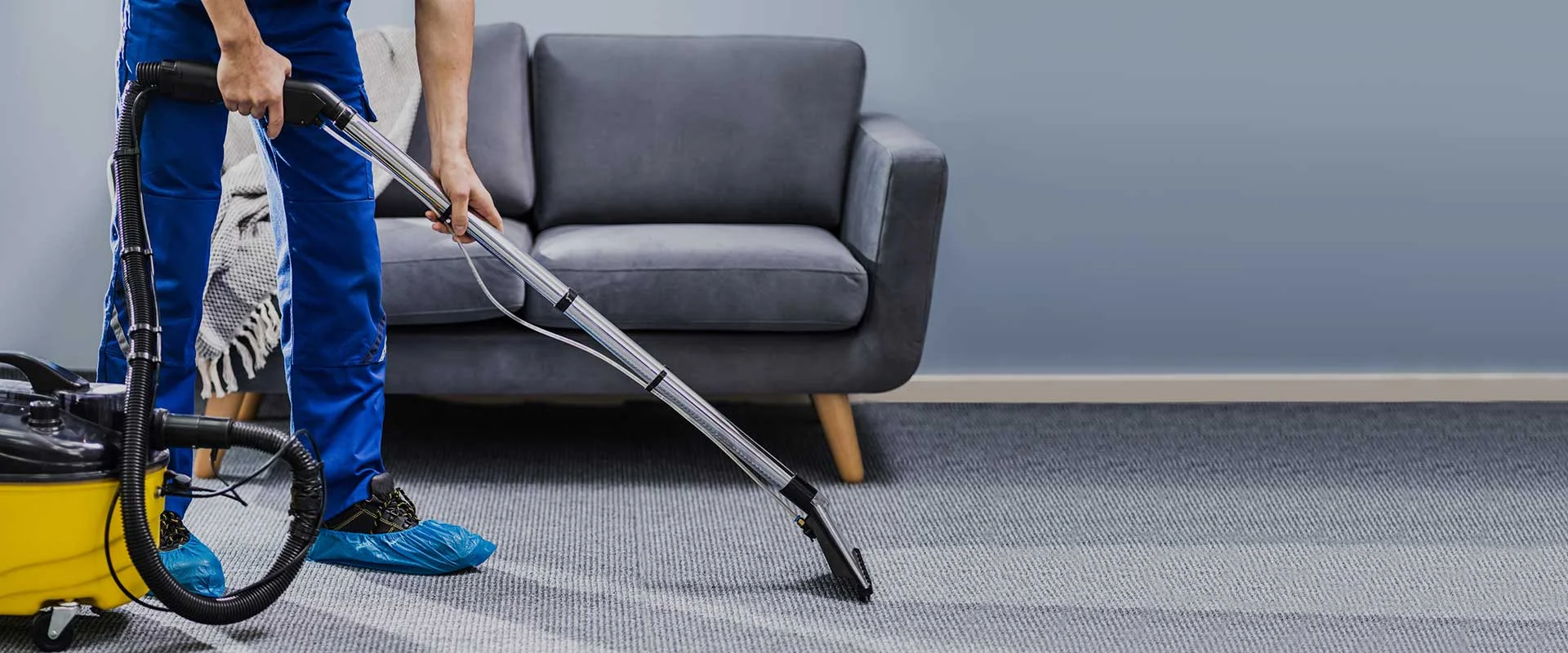 A person in blue overalls vacuum cleans a gray carpet with a yellow machine in a modern living room, next to a gray sofa.