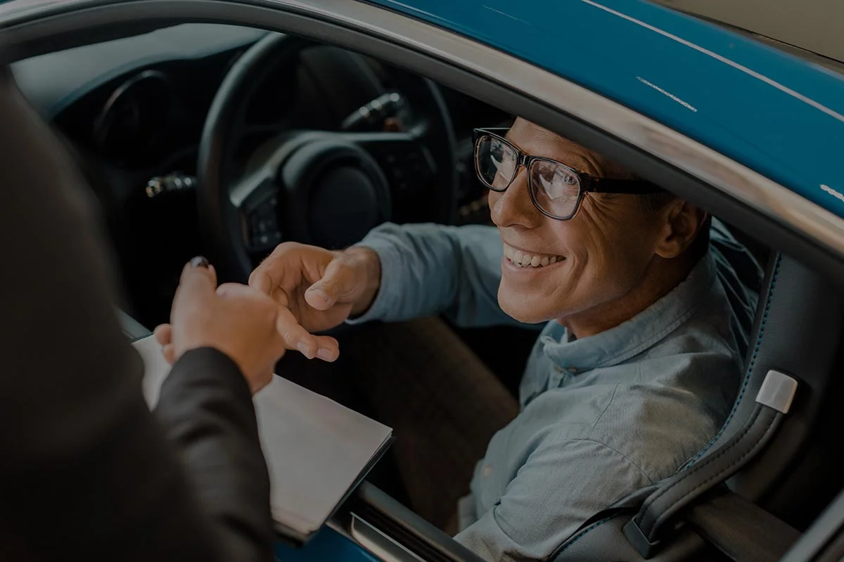 A smiling man wearing glasses sits in the driver’s seat of a blue car, reaching out to receive keys or documents from a person standing outside the vehicle.