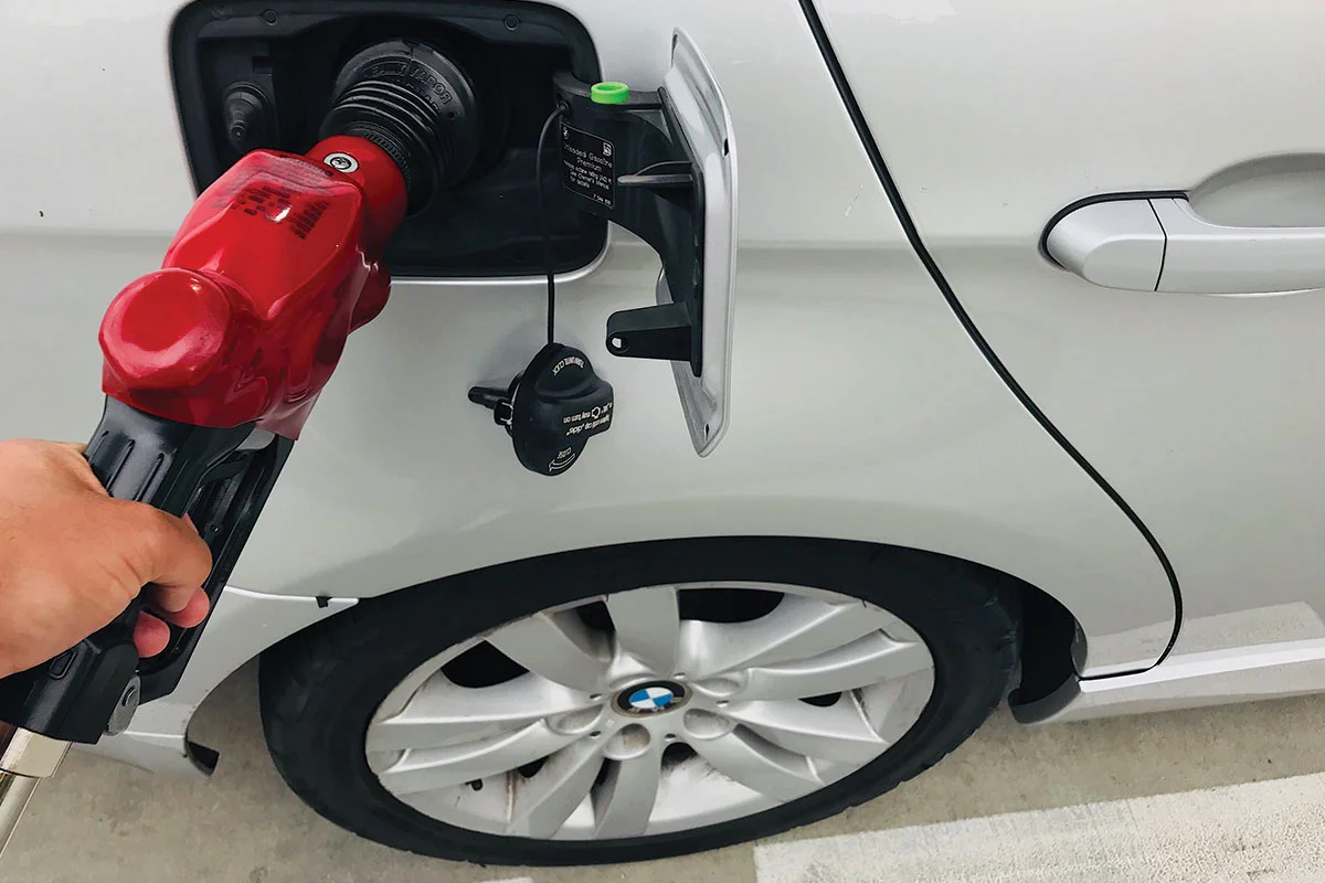 A person is holding a red fuel pump nozzle, refueling a white car at a gas station. The vehicle’s fuel cap is open, and the car’s rear wheel and BMW logo are visible.