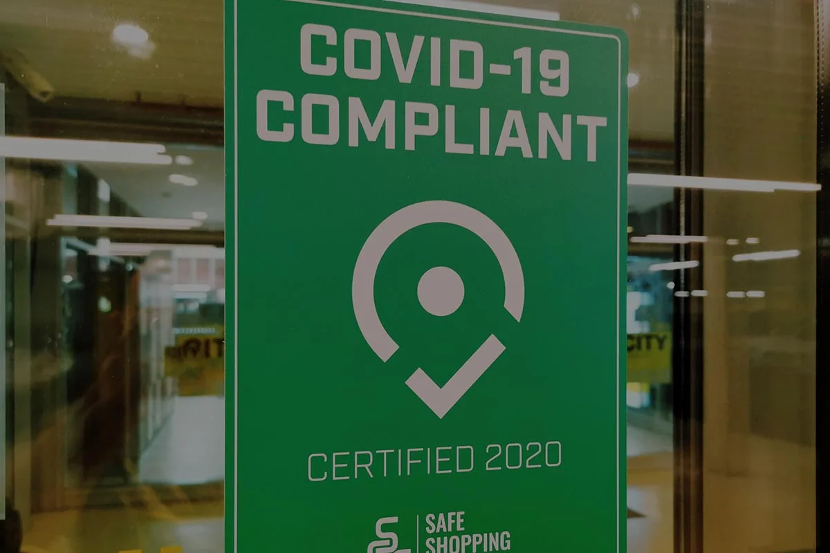 Green sign on a glass door reads "COVID-19 Compliant, Certified 2020" with a white circular icon and the words "Safe Shopping." Reflections of indoor lights and windows are visible in the background.