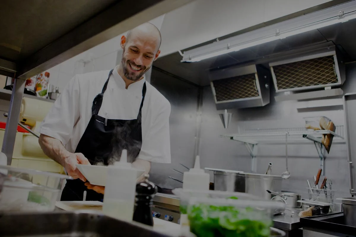 A chef wearing a white shirt and black apron smiles while preparing food in a professional kitchen, surrounded by cooking utensils, pots, and fresh ingredients.