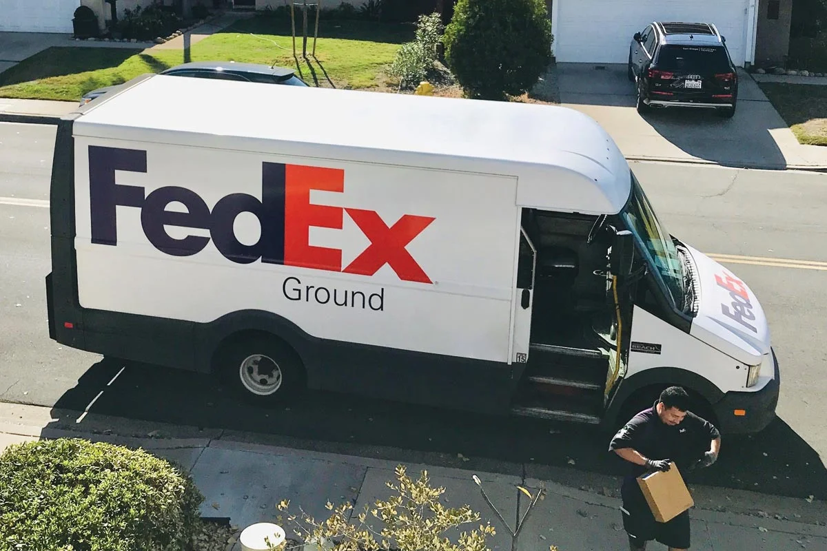A FedEx Ground delivery van is parked on a suburban street with its side door open. A delivery person is walking away from the van holding a package.