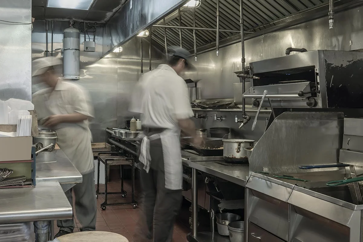 Two chefs in uniform and aprons work in a commercial kitchen with stainless steel appliances, stovetops, and pots; the scene appears busy with some motion blur.