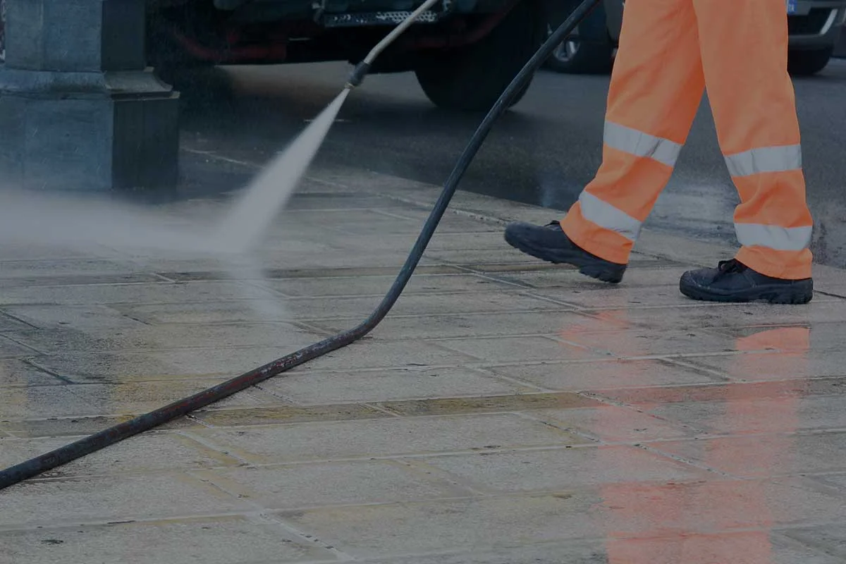 A person wearing orange reflective pants and black shoes is power washing a tiled sidewalk, with water spraying from the hose and wet tiles reflecting the scene.